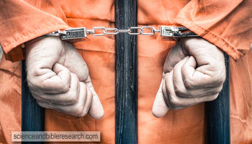 Handcuffed hands of a prisoner with orange clothes behind the bars - crispy desaturated dramatic filtered look (Photo by ViewApart)